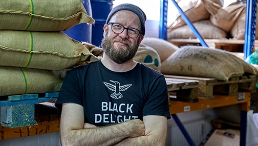 Neuigkeiten_Black-Delight_Ingo vor Kaffeesäcken im Rohkaffeelager der Black Delight Kaffeerösterei